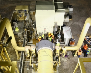 A group of engineers in hard hats work on a machine in a manufacturing facility.